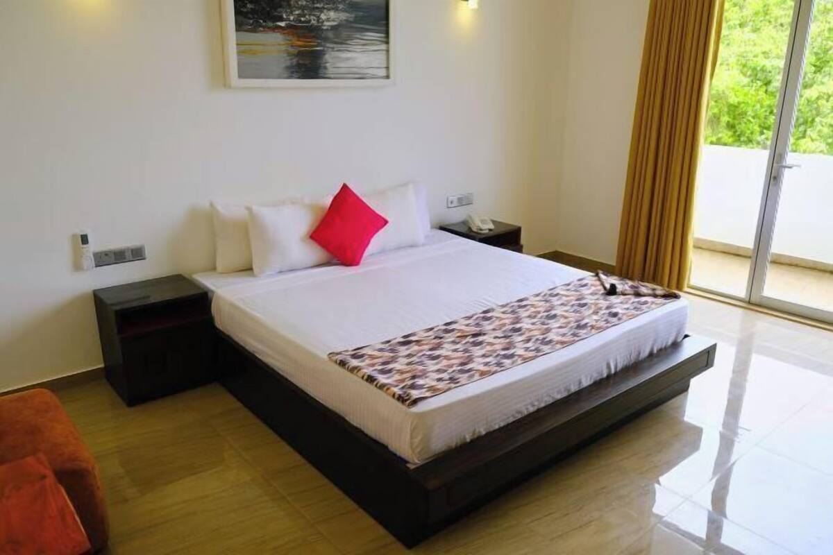 A modern hotel room with a large bed, a nightstand, a red pillow, and a view of greenery through the window.