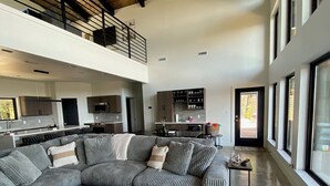 A spacious living area with a large sectional sofa, a kitchen in the background, and a high ceiling with exposed beams.