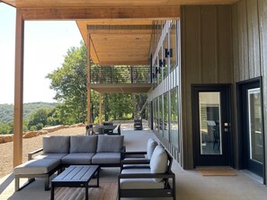 Et overdækket udendørs opholdsområde med moderne design, med en sofa og borde, og udsigt til et skovlandskab.