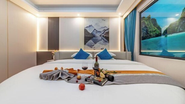A modern hotel room with a large bed, a painting of mountains, a scenic view through the window, and a table with a bottle and snacks. - Junyu Hotel (Guangdong Women's Vocational and Technical College Asian Games Village Branch) (Guangzhou)