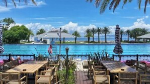 Zona junto a la piscina con mesas y sillas de madera, sombrillas y vista al océano.