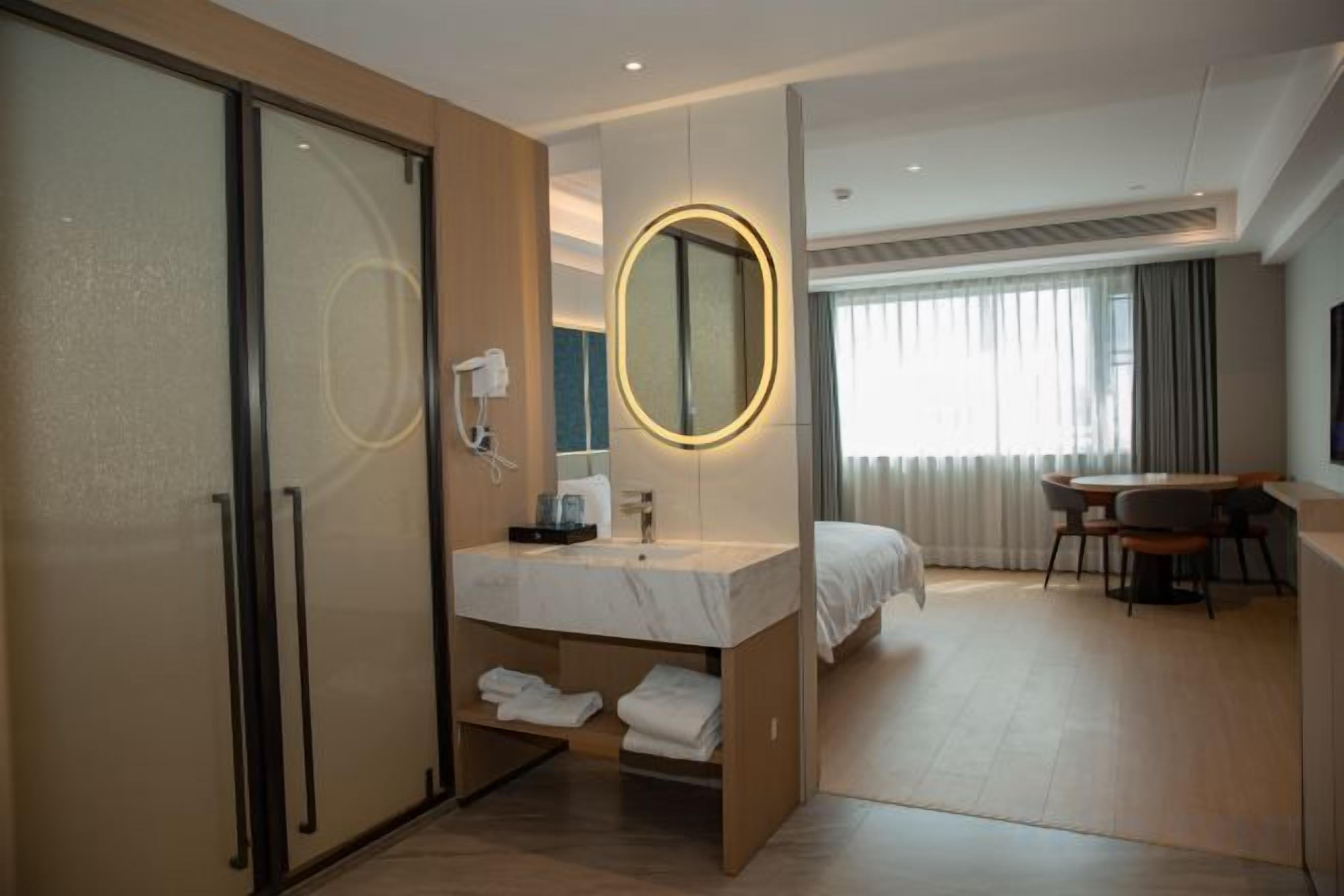 Habitación de hotel con armario correderas, baño con espejo y lavamanos, cama, mesa de comedor con sillas y un amplio ventanal con cortinas.