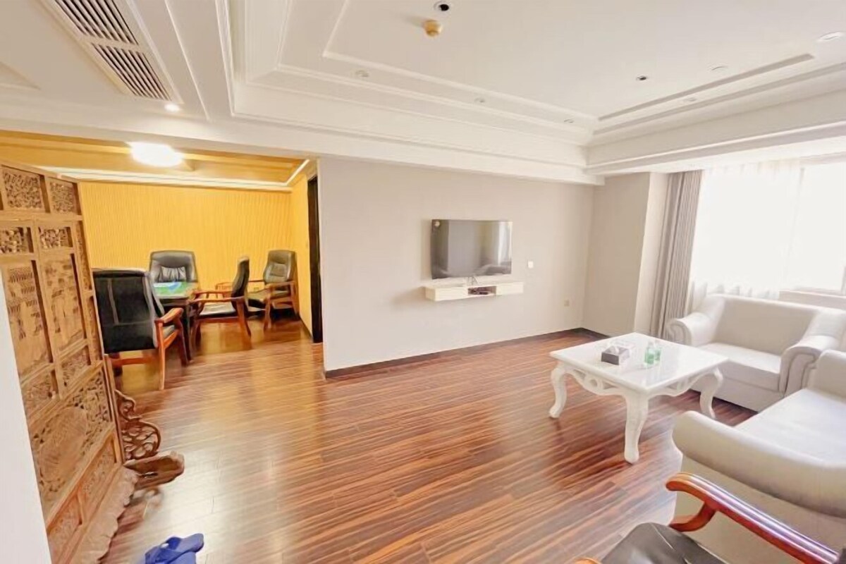 Ein Hotelzimmer mit Holzboden, einem weißen Sofa, einem weißen Sessel, einem kleinen weißen Tisch und einem an der Wand befestigten Fernseher.