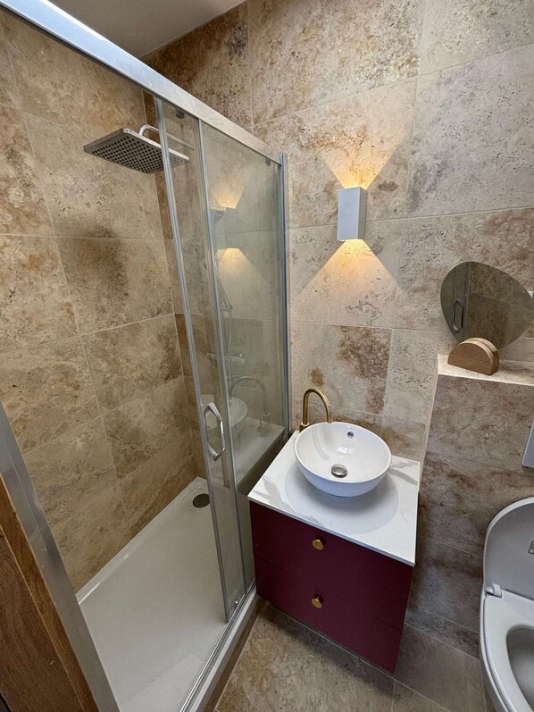 Un baño moderno con ducha envuelta en vidrio, un lavamanos blanco con grifo dorado y pared revestida de mármol.