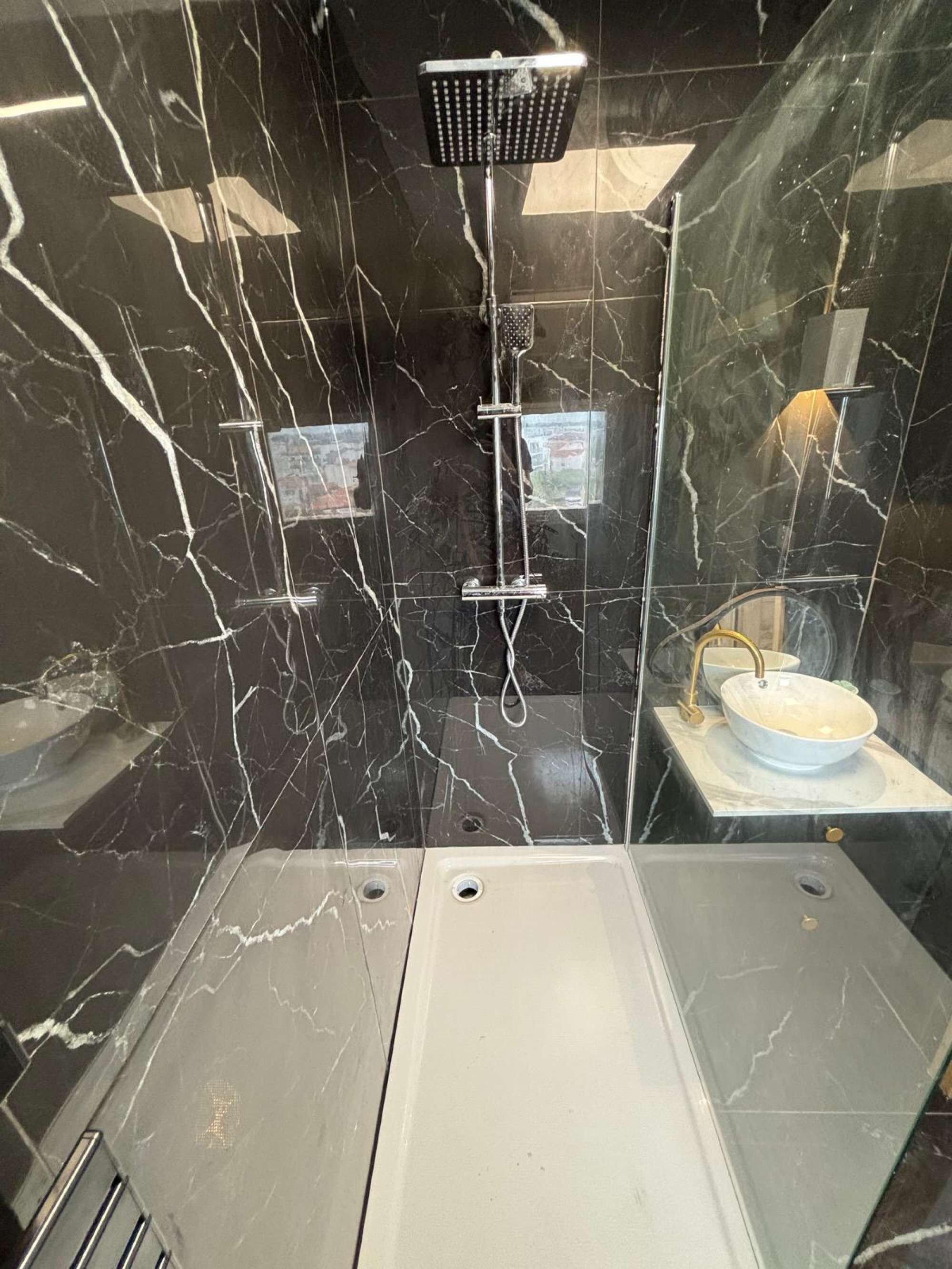 A modern bathroom with a black marble wall, a handheld showerhead, a sink with a gold faucet, and a mirror.