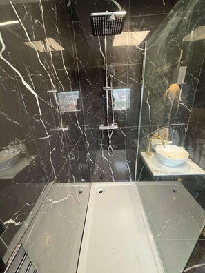 Un baño moderno con pared de mármol negro, cabezal de ducha de mano, lavamanos con grifo dorado y un espejo.