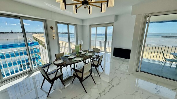Un comedor con una mesa dispuesta para cuatro personas, ventanas amplias que ofrecen vistas a la playa y la piscina, y un ventilador de techo.