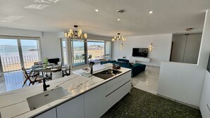Una cocina moderna con una isla grande, comedor con vistas a la playa y sala de estar con televisor y asientos.