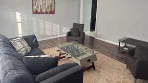 Una sala moderna con un sofá de cuero oscuro, una mesa de centro de cristal, un sillón gris y una mesita de noche de madera.
