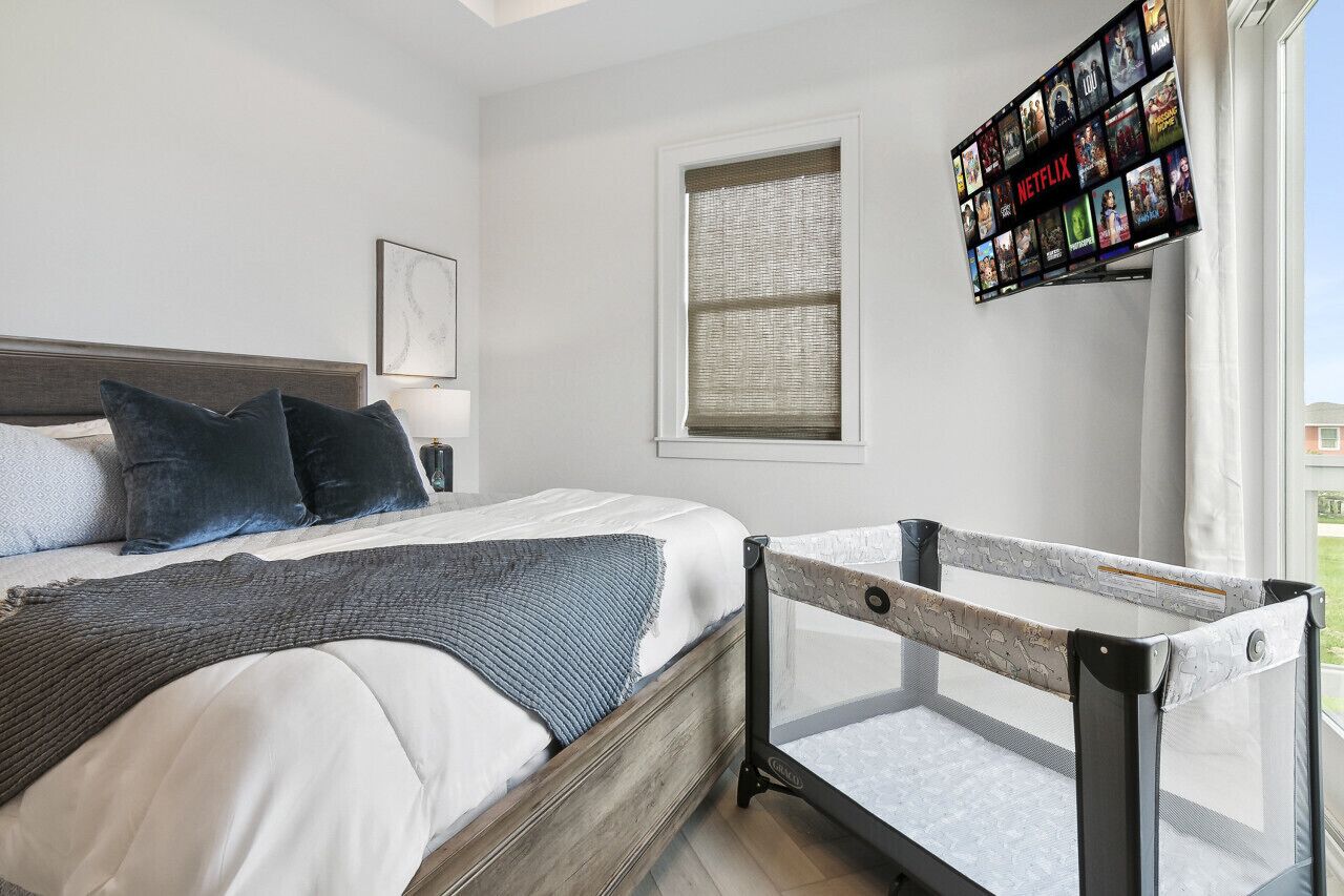 Ein Schlafzimmer mit einem Bett, einem Fenster mit Jalousien, einem an der Wand montierten Fernseher, der Netflix-Inhalte zeigt, und einem Babybett mit Sicherheitsgitter.