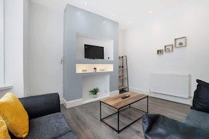 Ein modernes Wohnzimmer mit grauem Sofa, einem Holztisch, einem Wand-Fernseher und einer Heizung.