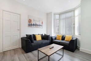 Un salon moderne avec un ensemble de canapés noirs, une table basse en bois et un tableau représentant un paysage urbain accroché au mur.
