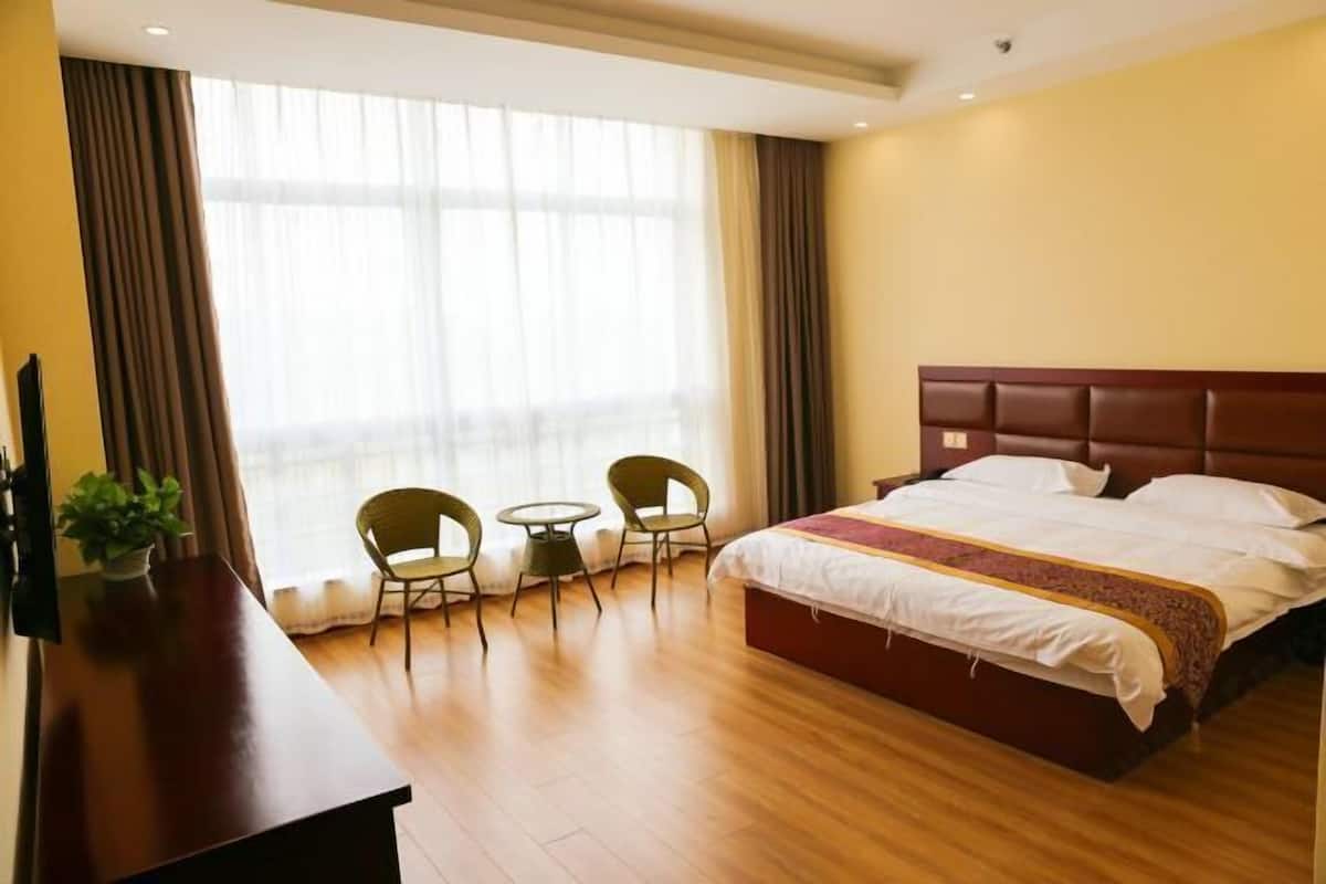 Une chambre d’hôtel avec un lit, un bureau, deux chaises, une télévision et une grande fenêtre avec des rideaux.