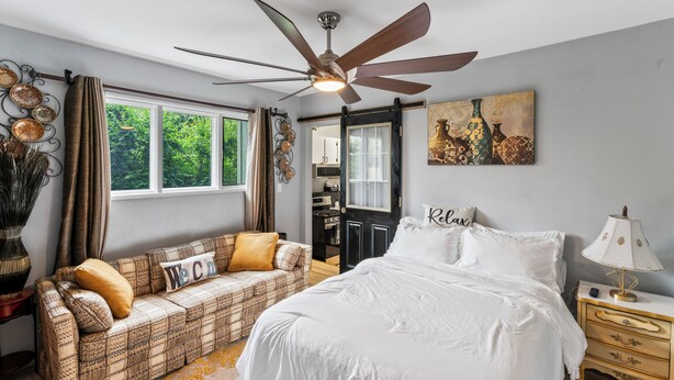 A bedroom with a bed, a sofa, a ceiling fan, a nightstand, a lamp, and a window with curtains. - Stylish Studio Suite Near Downtown Anchorage - Explore Alaska's Beauty with Ease (Anchorage)