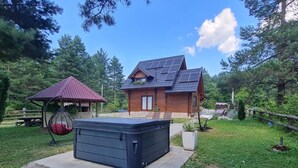 A wooden cabin with a sloped roof and solar panels, a hot tub, and a swing chair in a wooded area.