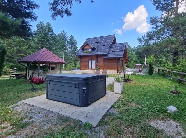 Une cabane en bois avec un toit en pente et des panneaux solaires, un jacuzzi et une chaise suspendue, le tout situé dans un espace boisé.