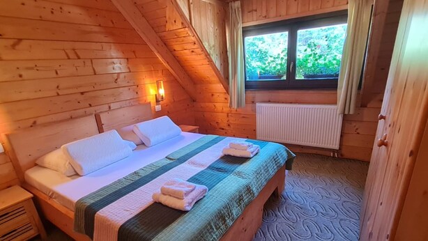 A wooden cabin bedroom with a bed, a nightstand, a window with curtains, and a radiator.