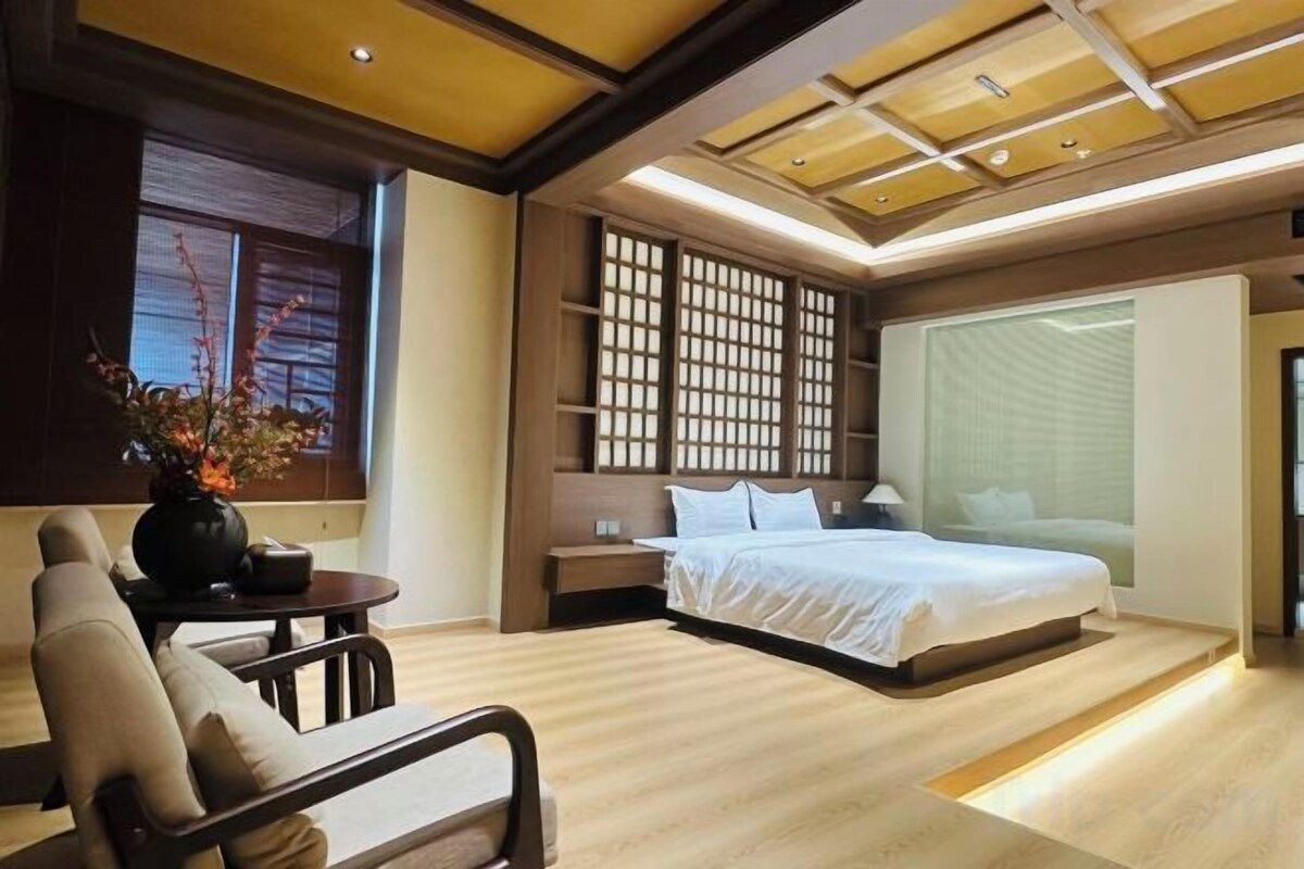 A modern bedroom with a large bed, bedside tables, a nightstand, a seating area with a chair, and a decorative vase.