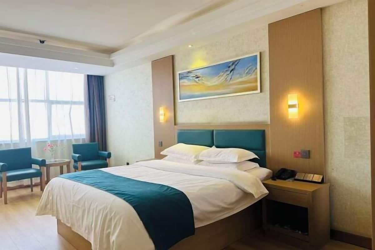 Une chambre d’hôtel avec un lit, deux chaises, un tableau, une table de chevet et une fenêtre avec des rideaux.