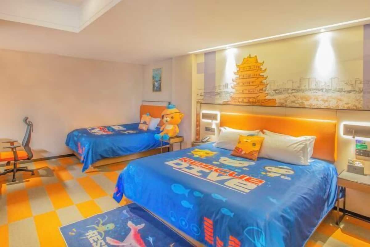 Une chambre d’hôtel avec deux lits, chacun recouvert d’une courtepointe bleue sur le thème du sauvetage. On y trouve un bureau avec une chaise, une lampe sur pied et un téléphone fixé au mur.