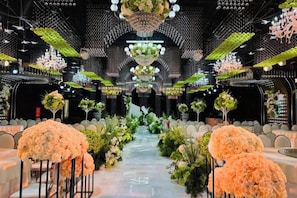 Un espace doté d’une allée centrale bordée de rangées de tables, chacune agrémentée de compositions florales et de chaises.