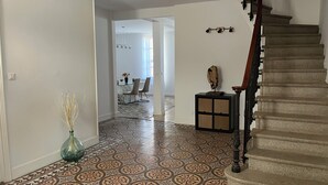 A hallway with patterned flooring leading to a dining area with chairs and a table. A vase with dried plants is on the left, and a small cabinet with a decorative item is on the right.