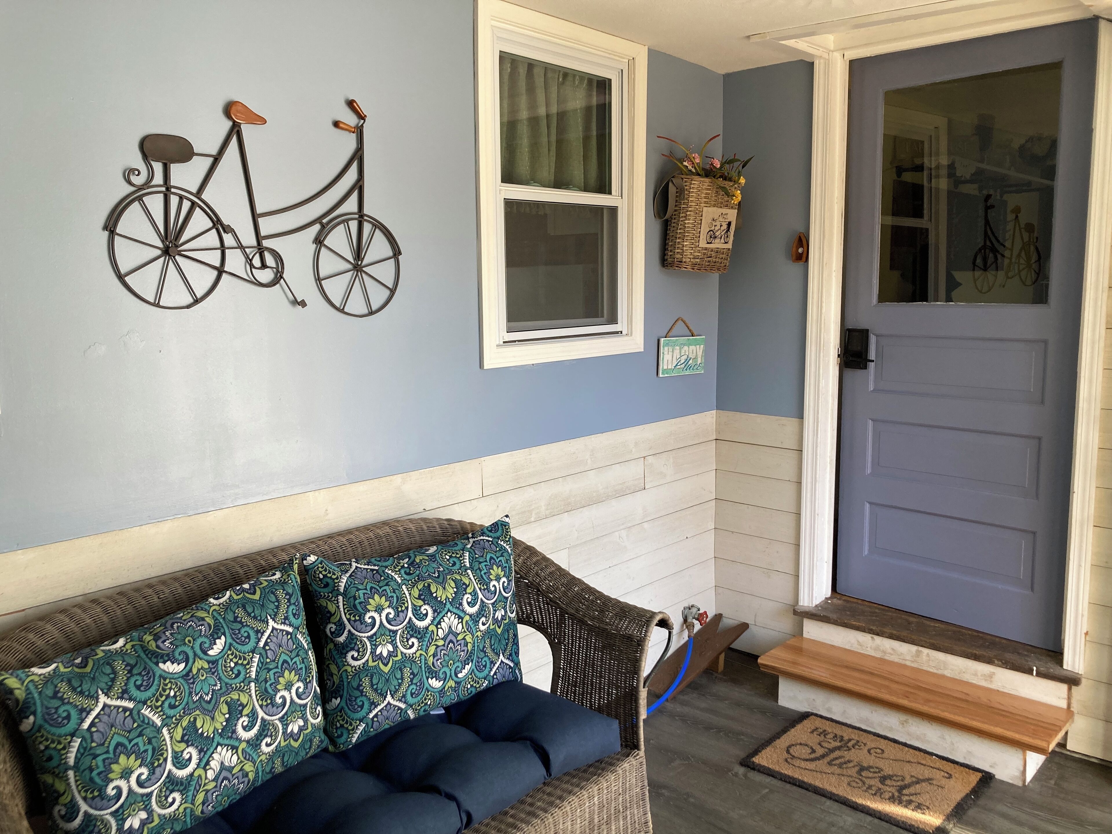 A cozy entryway with a wicker sofa, a wall-mounted bicycle decoration, a basket with flowers, and a door with a doormat.