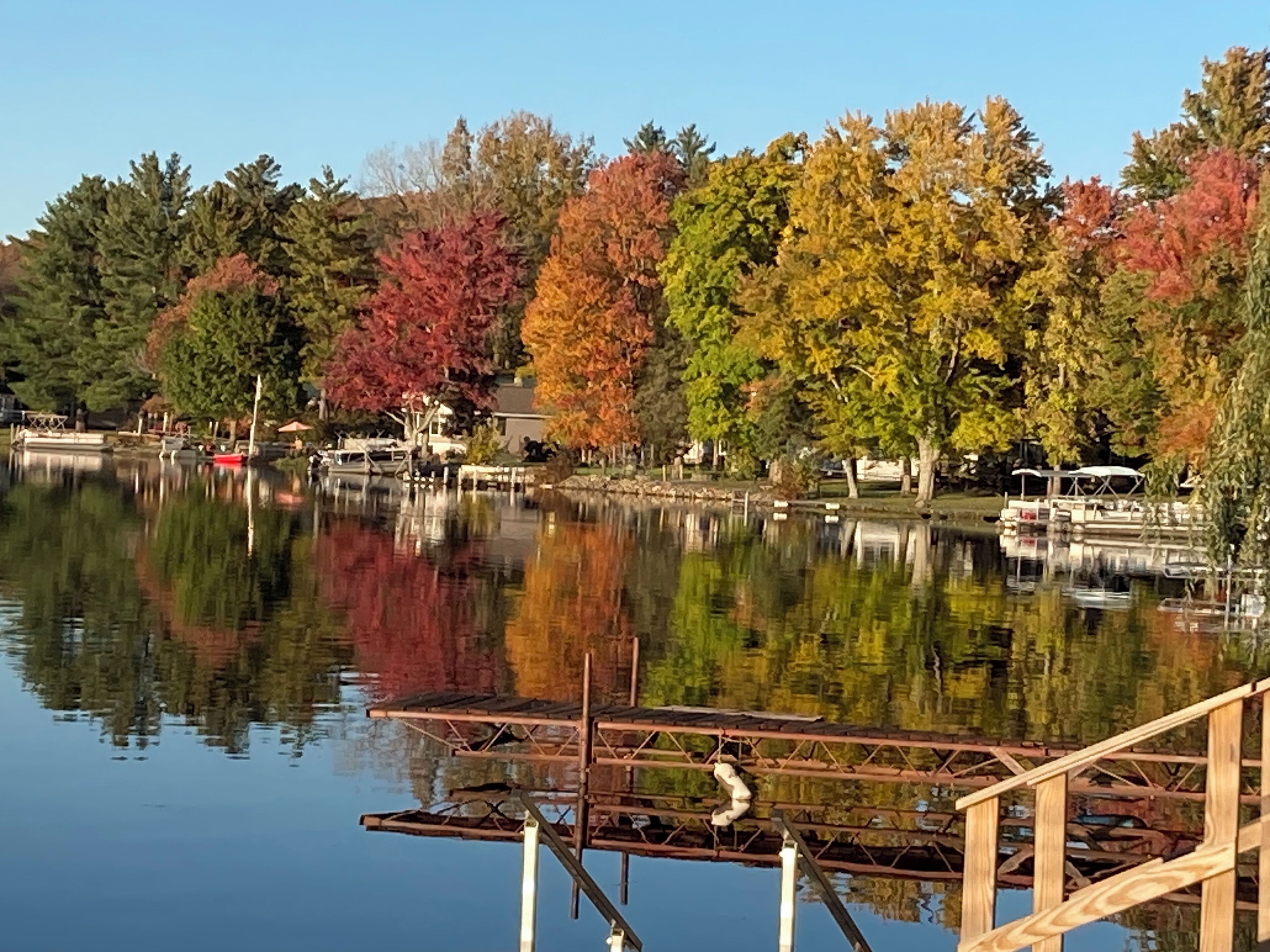 A calm lake with a wooden dock and boats, surrounded by trees with autumn foliage.