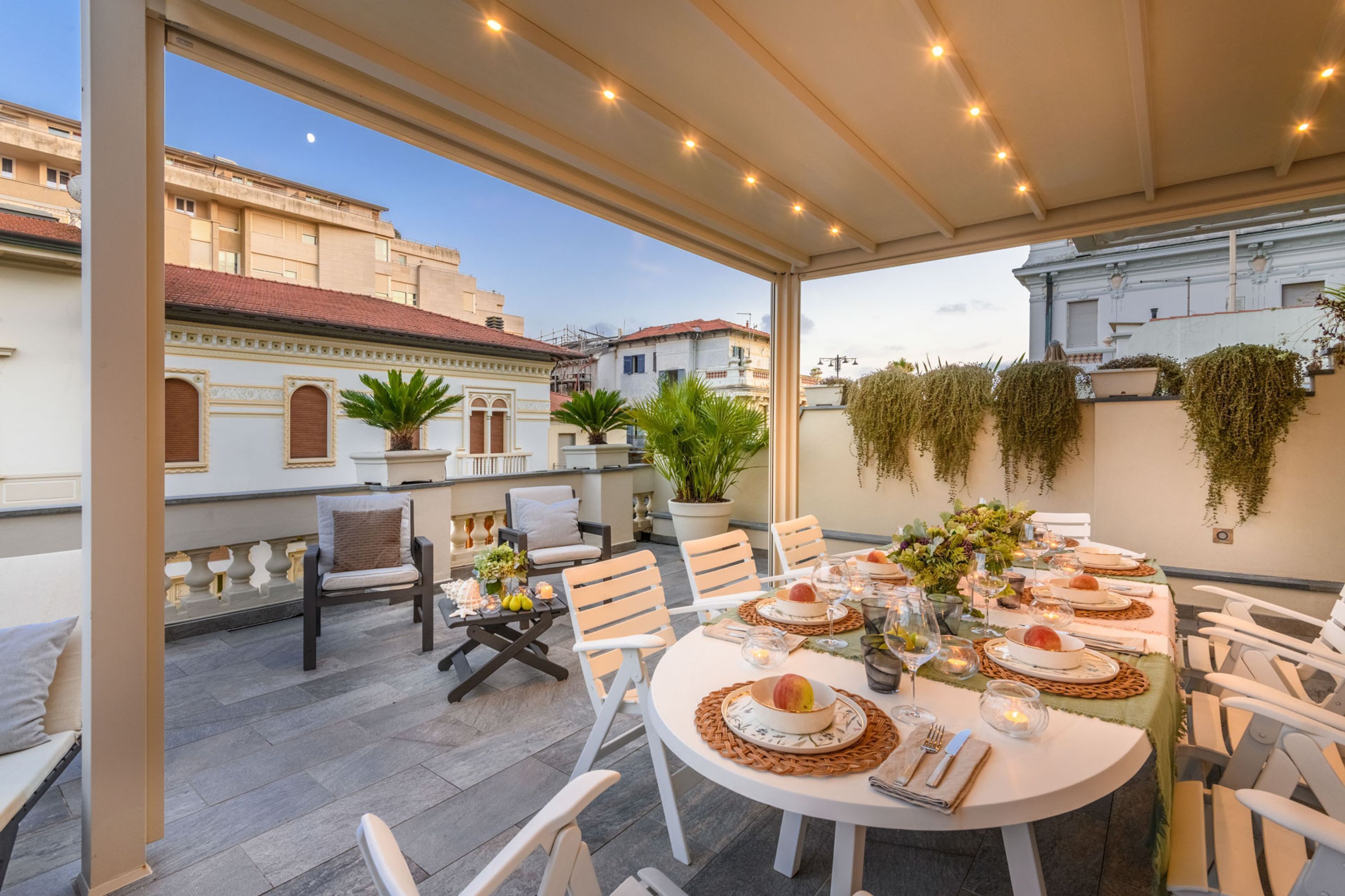A rooftop terrace with a round dining table set for six, surrounded by white chairs. There are potted plants and a hanging garden on the walls.