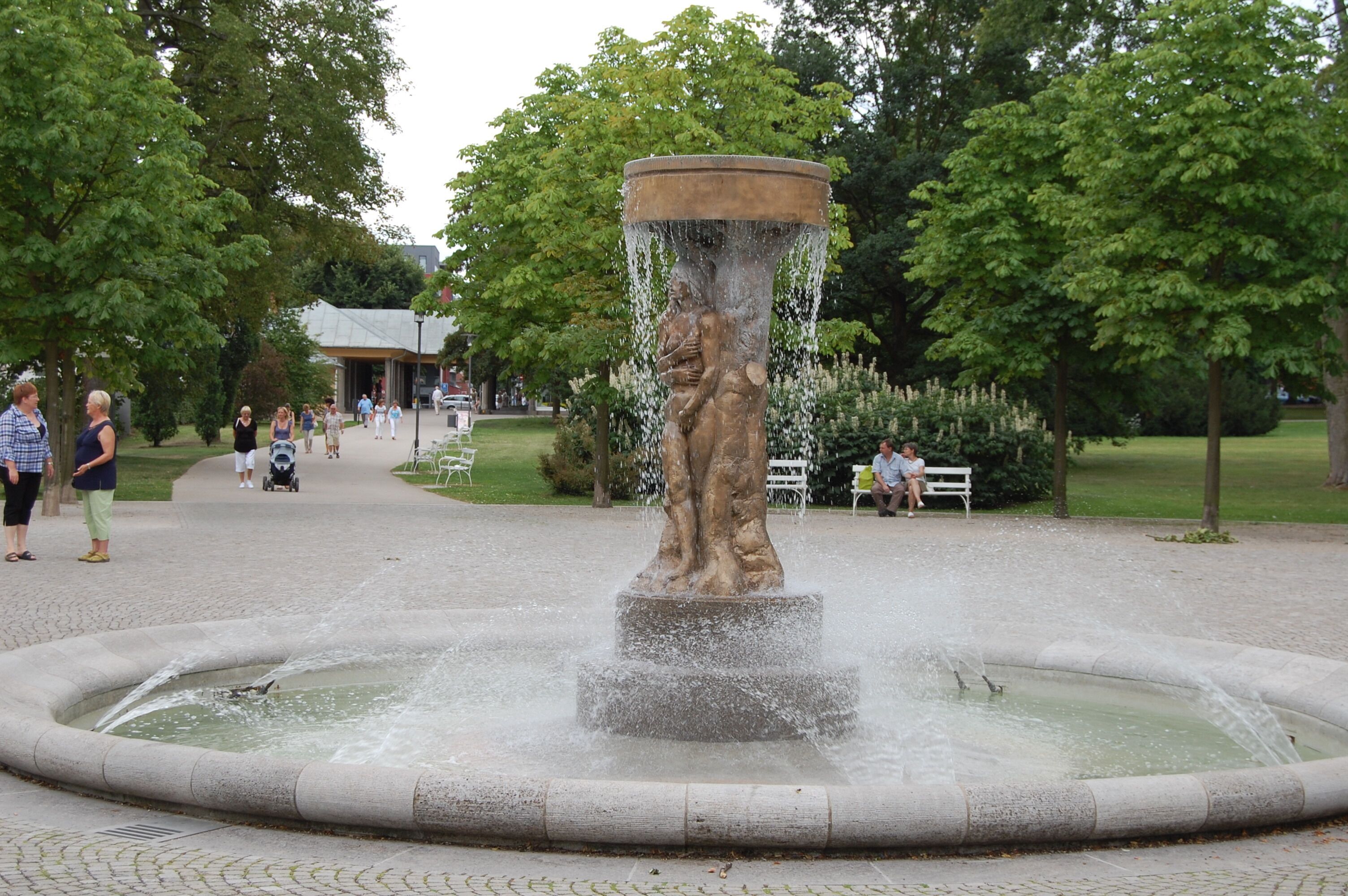 A fountain with water spouting from a central column, surrounded by a paved area with benches and trees.