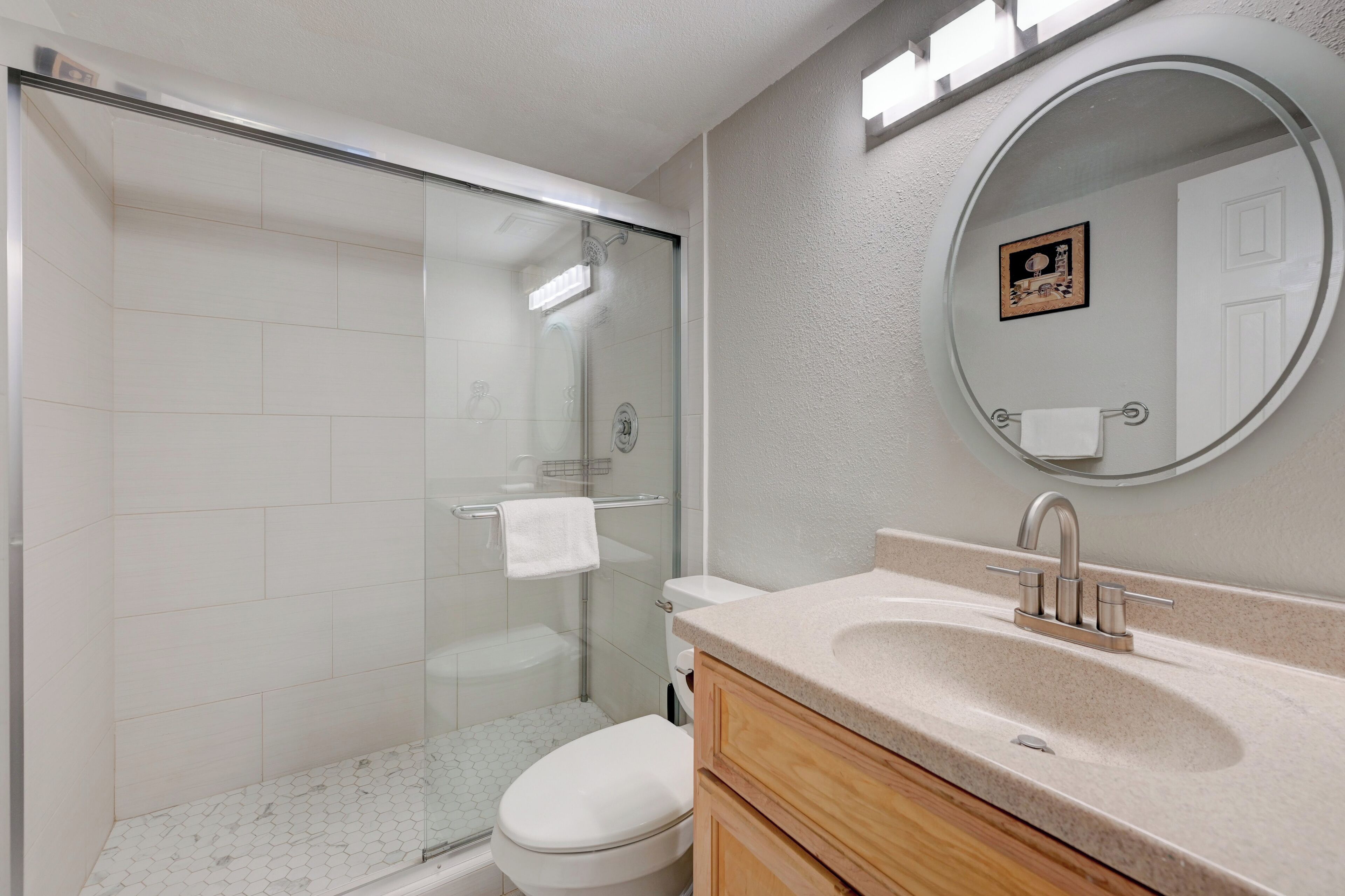 A modern bathroom with a glass-enclosed shower, a white toilet, a beige sink with a single faucet, and a round mirror with a picture frame.
