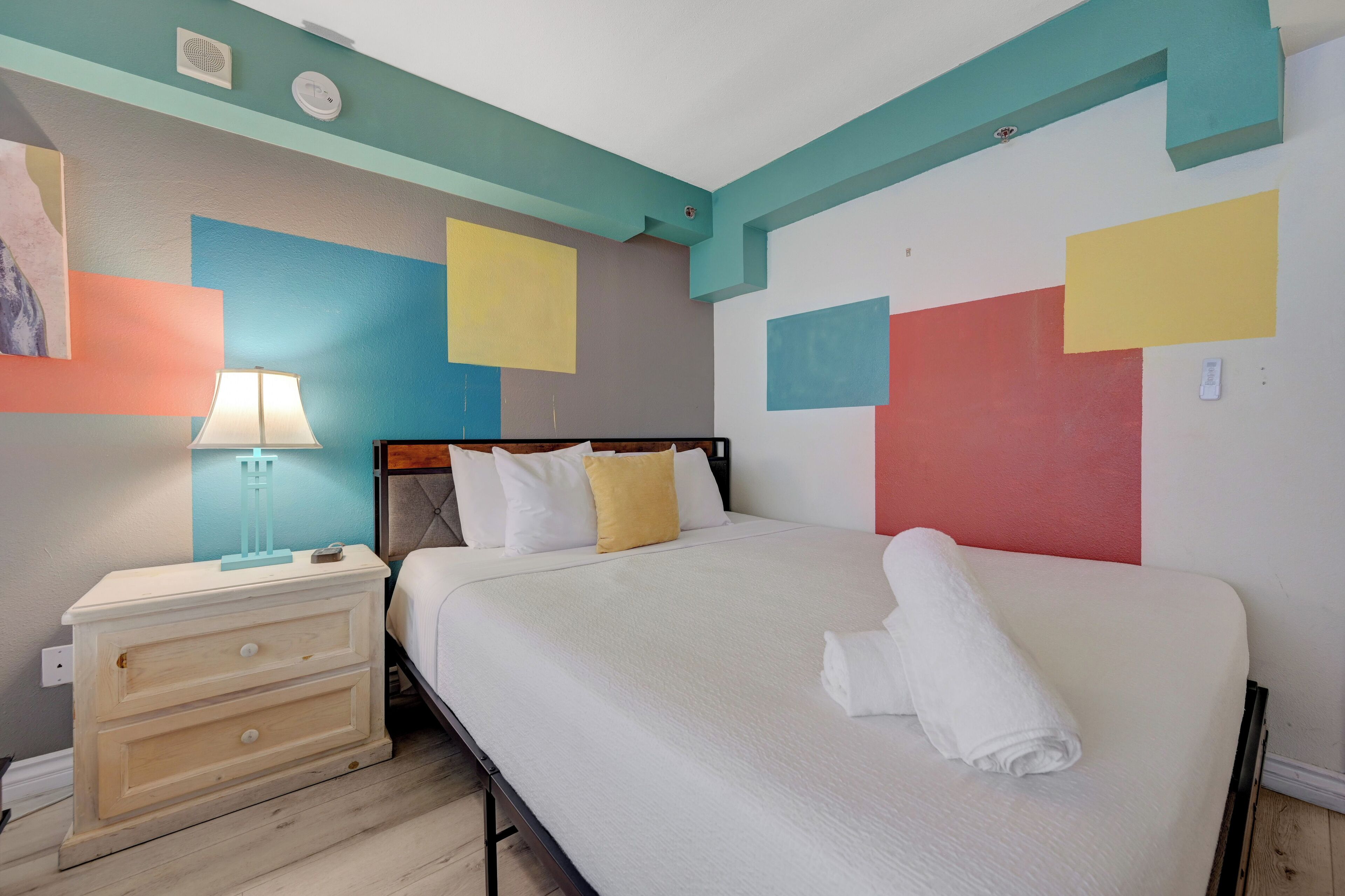 Un dormitorio con cama, mesita de noche y pared con formas geométricas coloridas.