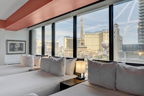 A hotel room with two beds, a nightstand with a lamp, and a large window offering a view of the Eiffel Tower and other city landmarks. - Stay Together Suites | Center Strip PH (Las Vegas)