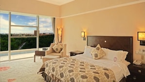 A hotel room with a large bed, a chair, a nightstand, a lamp, and a view of greenery through a large window. - University Town International Hotel (Guangzhou)