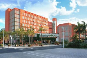 Un hotel de varios pisos con una fachada roja llamativa, un acceso cubierto y un área ajardinada con palmeras.