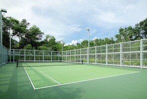 An indoor tennis court with a green surface, surrounded by a metal fence, and trees in the background.