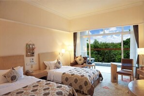 Habitación de hotel con dos camas, una mesita, una silla y un balcón con vistas a zonas verdes.