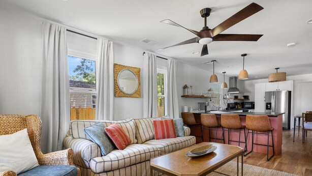 Een moderne woonkamer met een bank, fauteuils en een eettafel. In de achtergrond is een keuken met een bar en een plafondventilator zichtbaar.
