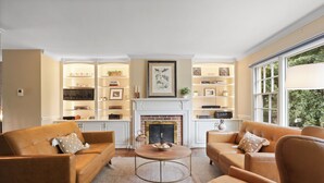 A living room with two sofas, a coffee table, and a fireplace. There are shelves with decorative items and a window with a view.