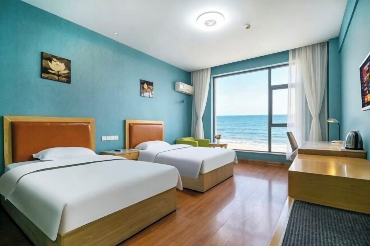 Quarto de hotel com duas camas, uma escrivaninha, uma cadeira, vista para o mar e uma janela com cortinas.
