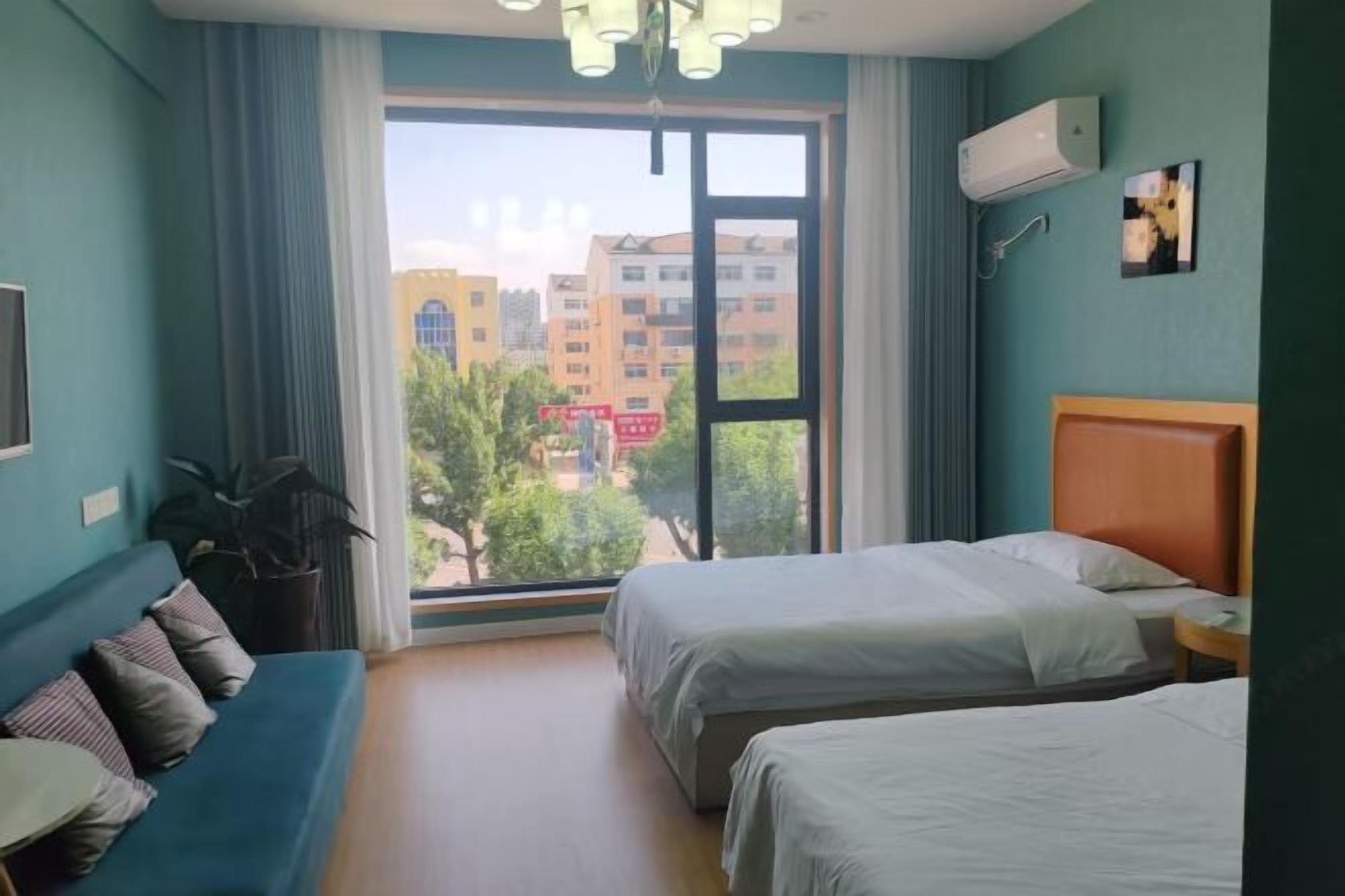 A hotel room with two beds, a sofa, a large window with a view of buildings, and an air conditioning unit.
