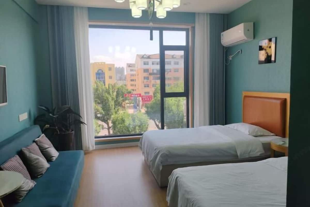A hotel room with two beds, a sofa, a large window with a view of buildings, and an air conditioning unit.