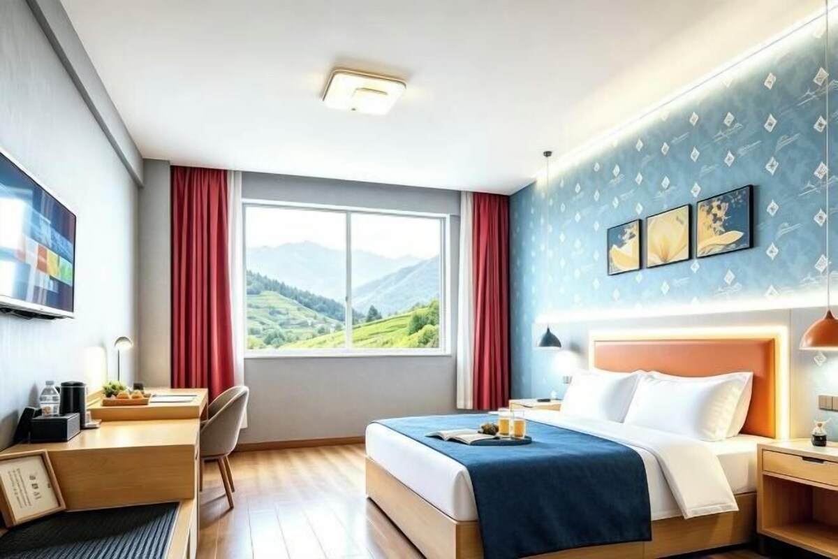 Quarto de hotel com cama, escrivaninha, cadeira e uma janela grande com vista para as montanhas.