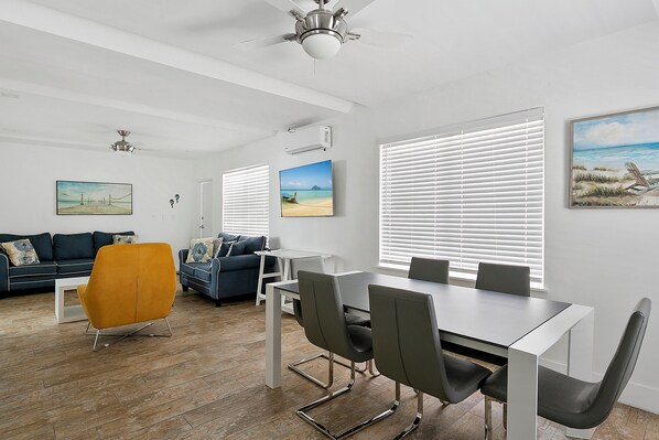A modern living room with a dining area, featuring a sofa, a coffee table, and a dining table with chairs. There are large windows with blinds, a ceiling fan, and artwork on the walls. - Spacious 1 Bedroom APT at the beach (Hollywood)