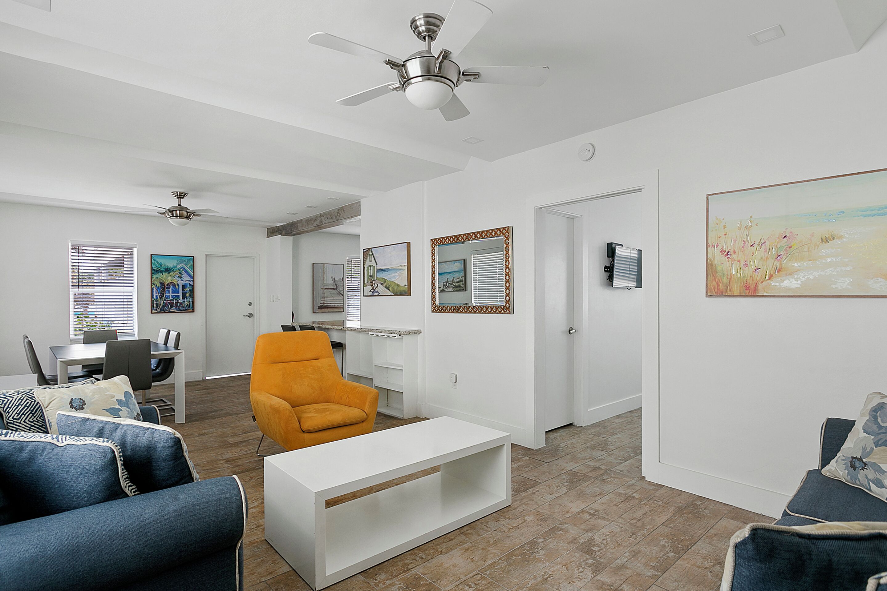 A spacious living room with a white sofa, a bright orange armchair, and a white coffee table. There are ceiling fans, a dining area with a table and chairs, and a hallway leading to another room.