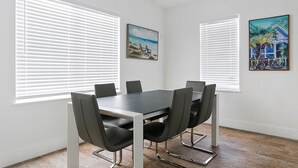 A modern dining room with a rectangular table and six chairs, large windows with blinds, and two framed paintings on the walls. - Spacious 1 Bedroom APT at the beach (Hollywood)