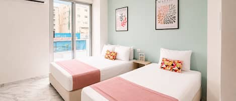 A hotel room with two beds, a window with a view of buildings, and two framed artworks on the wall.