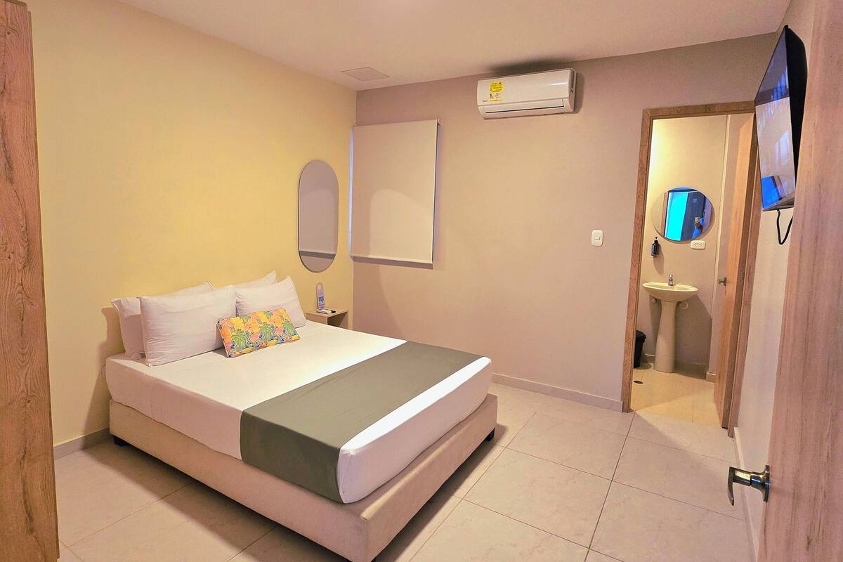 Une chambre à coucher comprenant un lit, une table de chevet, un miroir, un appareil de climatisation et une salle de bain visible à travers une porte ouverte.