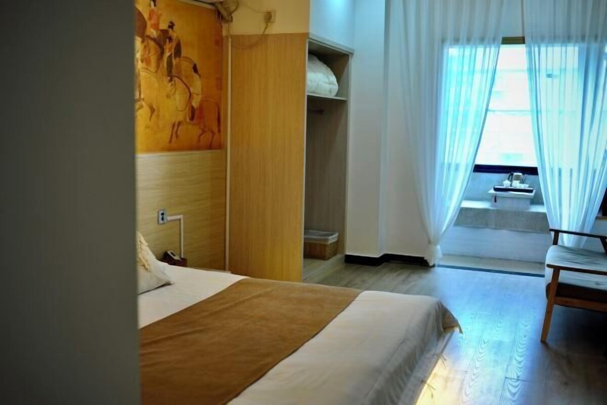 A hotel room with a bed, a bedside table, a chair, a wardrobe, a painting on the wall, and a window with curtains.