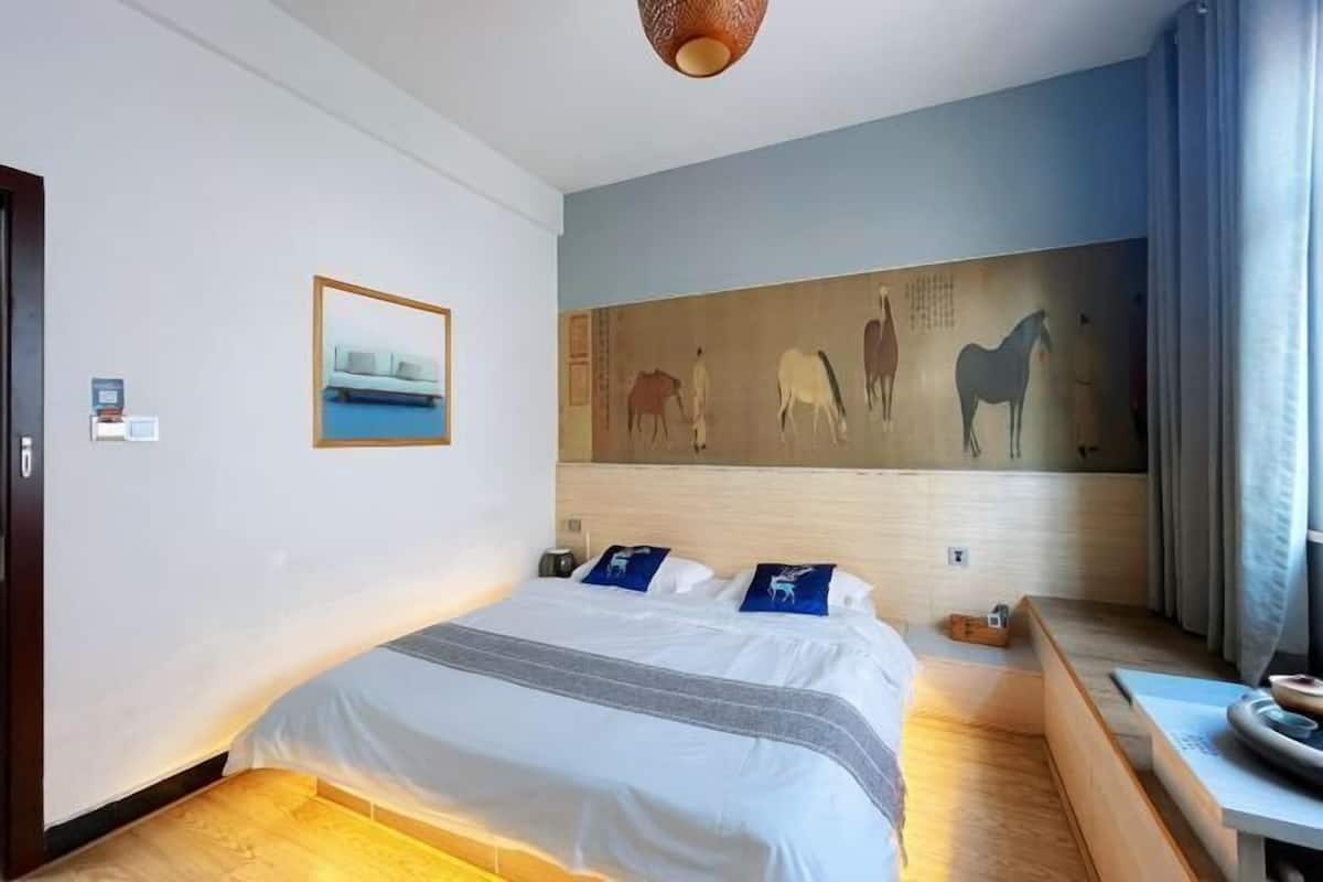 A hotel room with a bed, a painting of horses on the wall, a small table, and a window with curtains.